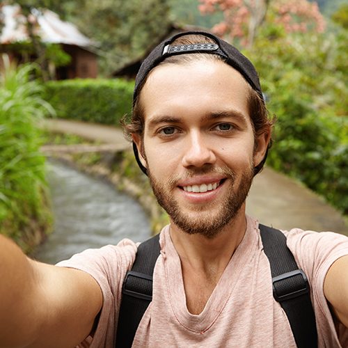 Close up view of happy face of attractive hiker with beard smiling while taking selfie, holding camera with both hands during his journey in countryside. Young hipster spending holidays in village