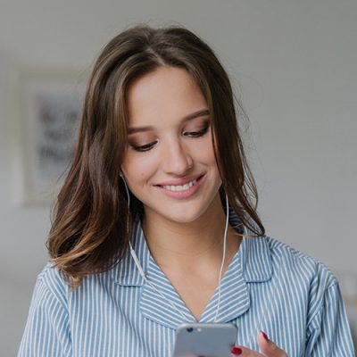 Happy woman with appealing look, holds modern cell phone, downloads song to playlist, listens music with earphones, sits crossed legs on comfortable bed, has charming smile on face, uses free wifi