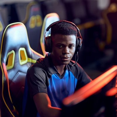 Portrait of a happy african guy, professional gamer wearing headphones looking at camera and smiling while participating in eSport tournament, sitting in gaming club or internet cafe